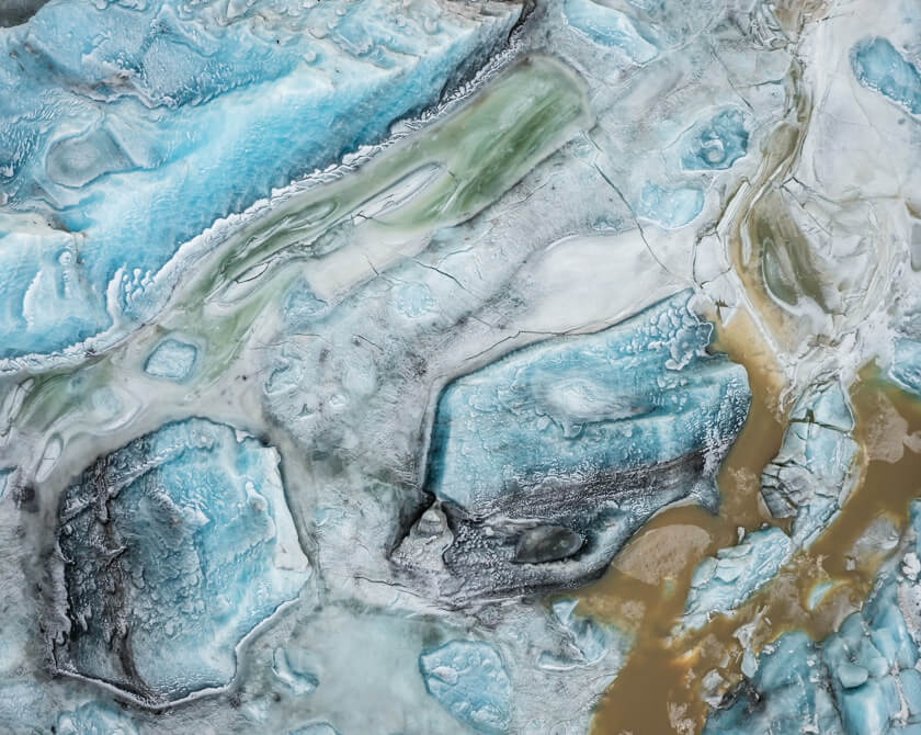 Drone perspective of Svínafellslón in winter, where ice formations reflect blues, greens, and earthy tones